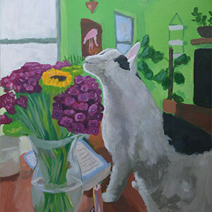 In a room with green walls, a white cat with black spots smells purple and yellow flowers in a clear vase.